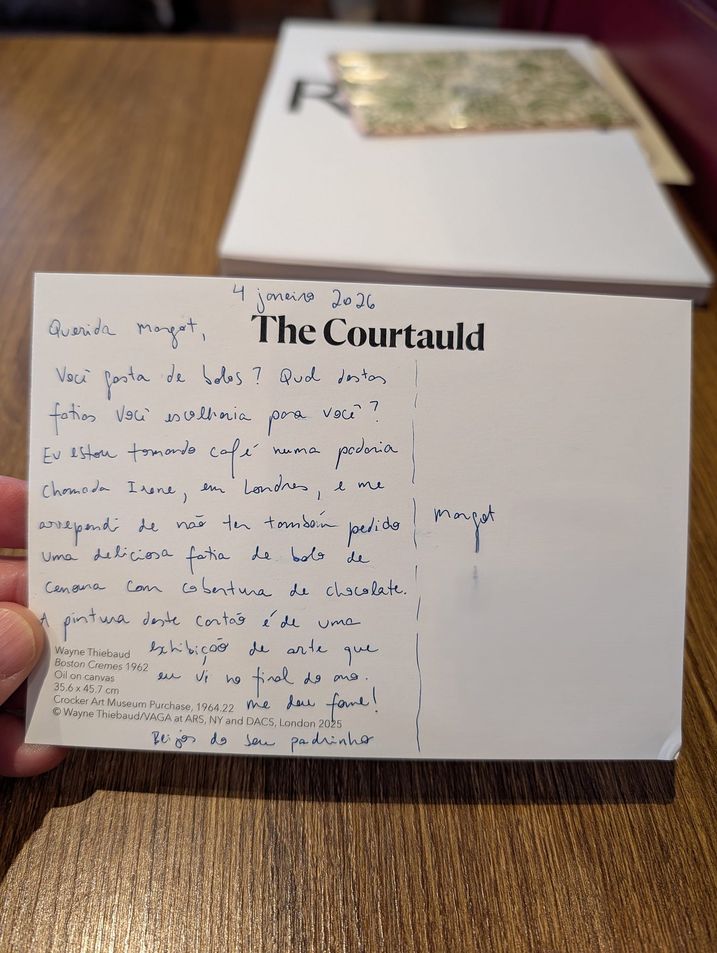 A postcard addressed to 'Querida Margot' (Dear Margot) dated 4 January 2026, written in Portuguese. The handwriting fills the left side of The Courtauld postcard, asking about cakes and mentioning coffee at a London bakery called Irene. The postcard sits on a dark wooden table with another card visible in the background.