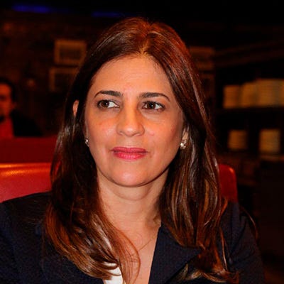 Mulher branca, cabelos e olhos castanhos, usa blazer preto e blusa branca. Fundo escuro.