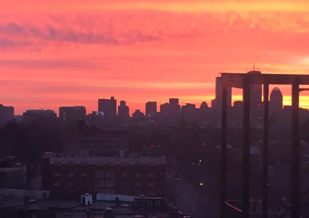 The silhoutte of the Boston skyline set against a pink and yellow sunrise.