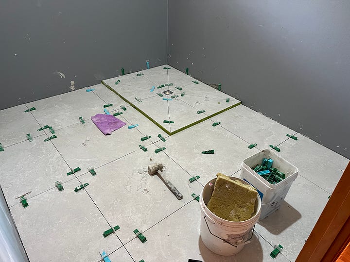 four photos, the first one shows a dated bathroom with dark floor tiles and a shower with a big monstera plant in front of it. The next photo shows the same space with new whitish grey floor tiles just laid. The third photo is an outdoor shot with a pile of garbage that includes: an old toilet, ripped up tiles and other debris from a housing demolition. The final photo is of a front porch with a yellow screen door in the center. On one side is a tile cutting machine on a drop cloth with many other tools of the trade and on the opposite side is a pile of wood trim and boxes of tiles.