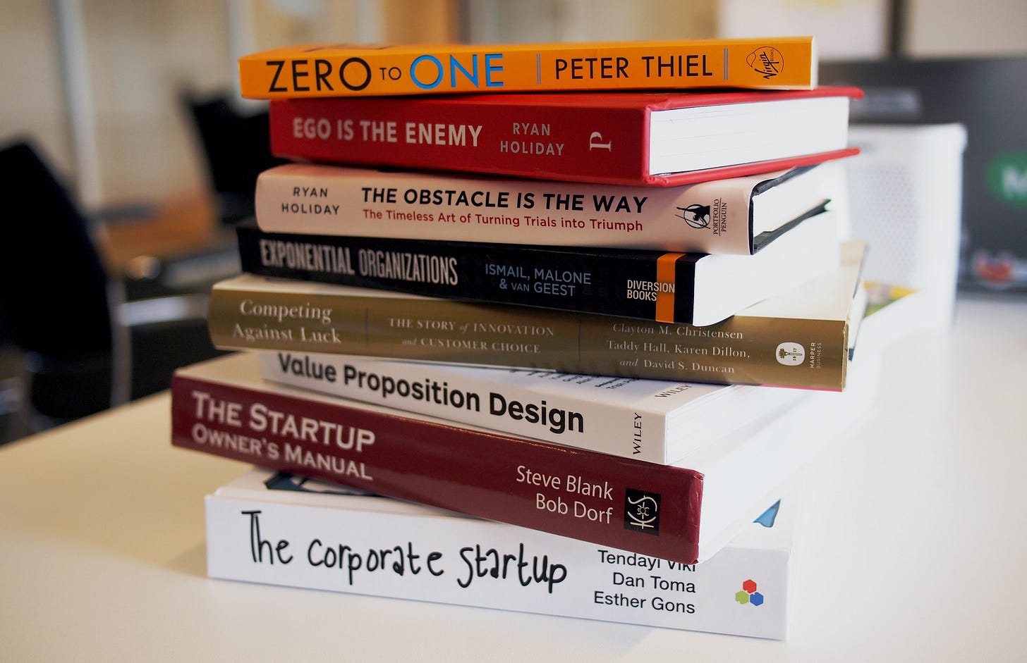 A stack of business books on a desk: Zero to One, Ego is the Enemy, Obstacle is the Way, Exponential Organizations, Competing Against Luck, Value Proposition Design, The Startup Owners Manual, The corporate startup. A stack of business books on a desk: Zero to One, Ego is the Enemy, Obstacle is the Way, Exponential Organizations, Competing Against Luck, Value Proposition Design, The Startup Owners Manual, The corporate startup.