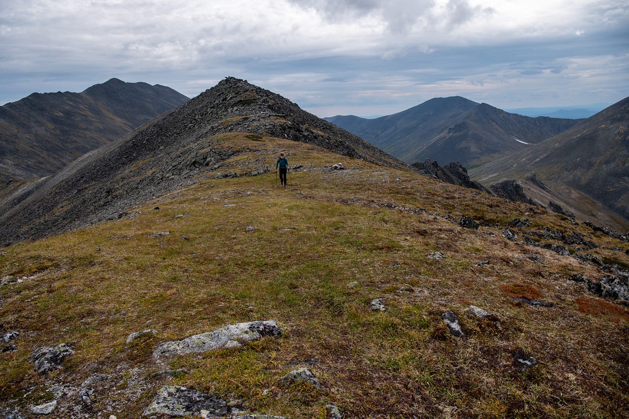 A lone hiker crosses a broad, grassy alpine ridge dotted with rock, walking toward the camera with peaks and rubble slopes falling away on both sides under a heavy grey overcast sky.