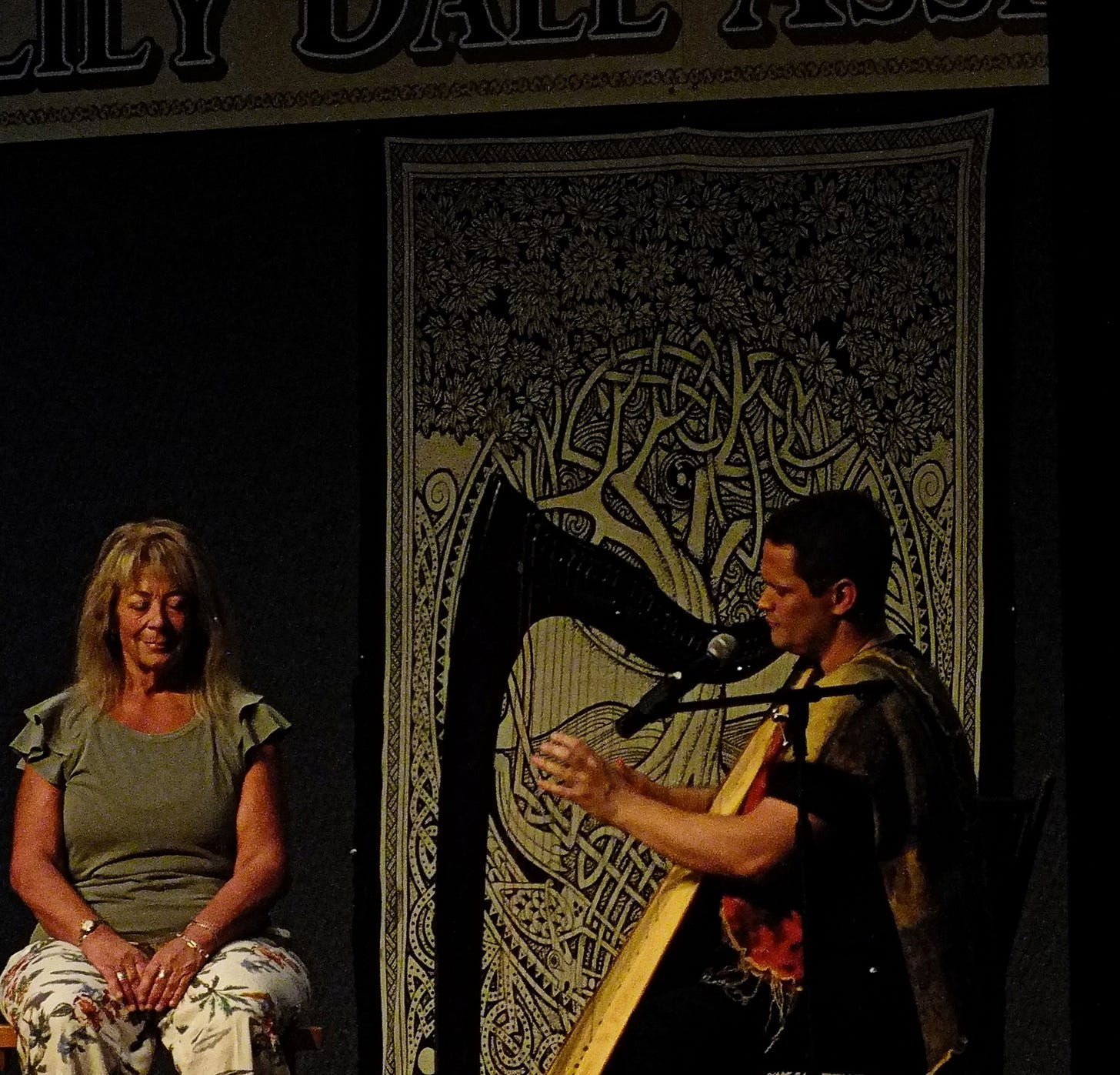 Russell playing the harp on stage in Lily Dale while a woman is seated and receiving the music intuited for her.