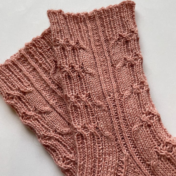 Two heavily cabled socks knit in pink yarn.