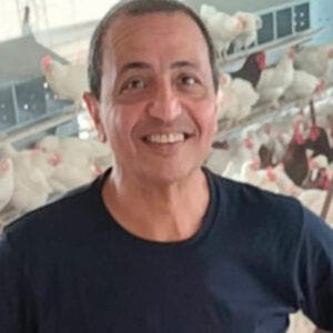 Homem sorridente de cabelos grisalhos usando camiseta azul tendo ao fundo um criadouro de frangos