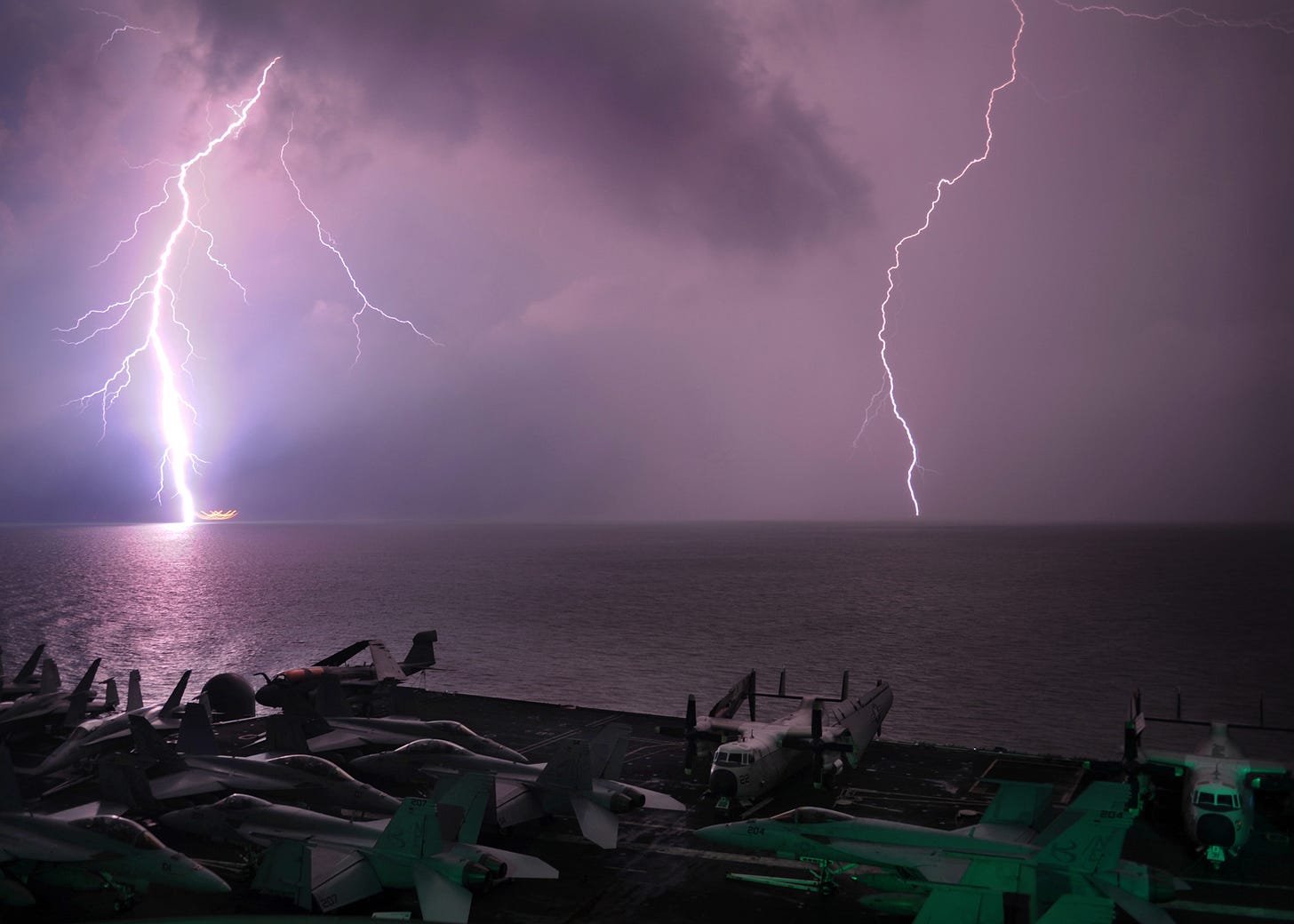 Lightning strikes over the ocean, seen from an aircraft carrier deck with several military planes visible in the foreground at night.