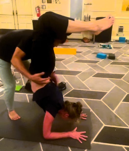 The author is shown on her yoga mat, upside down and in a backbend shape, in a shape known as Scorpion pose. There is a teacher assisting her to do the pose safely, but his hands are not supporting her directly in this image. The author is shown on her yoga mat, upside down and in a backbend shape, in a shape known as Scorpion pose. There is a teacher assisting her to do the pose safely, but his hands are not supporting her directly in this image.