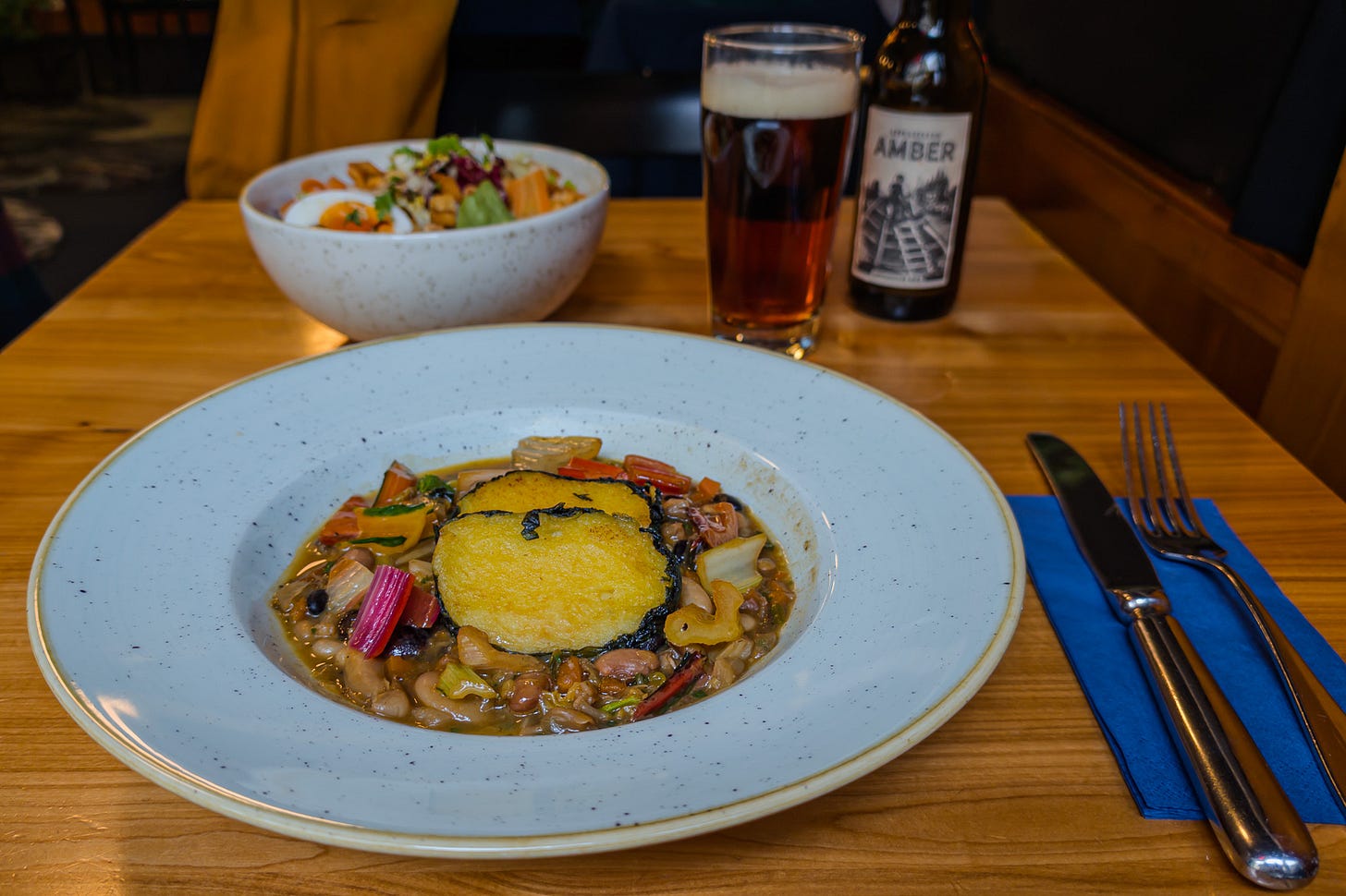 Vegetarisches Gericht auf einem grossen weissen Teller: gebratene Polenta, in Mangoldblätter gewickelt, auf einem Ragout aus Hülsenfrüchten. Im Hintergrund stehen eine weitere Gemüseschale sowie ein Glas und eine Flasche Amber-Bier.