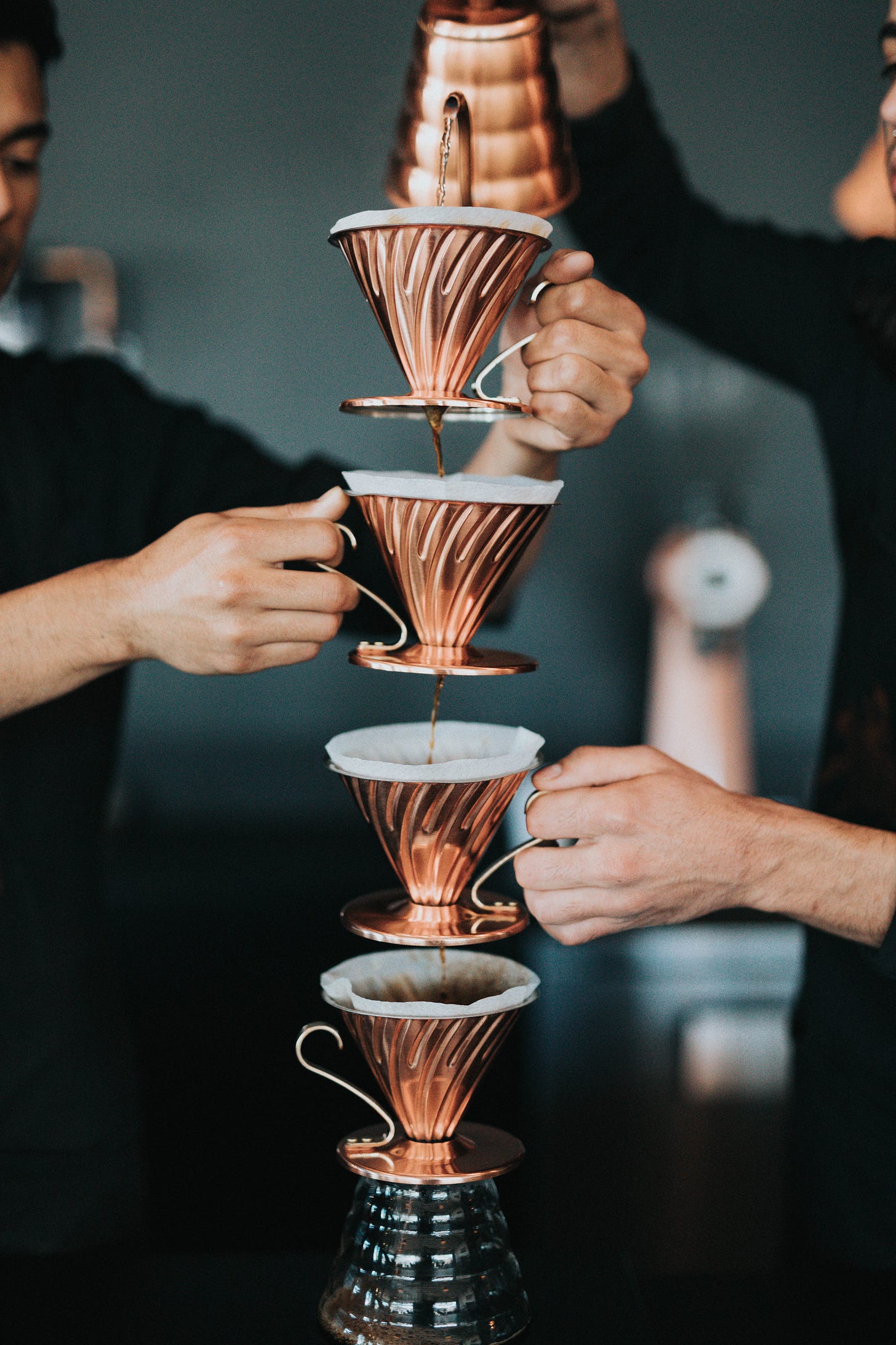 A couple baristas pouring hot water through 4 stacked V60s with paper filters, into a carafe. This is a very silly way of making coffee