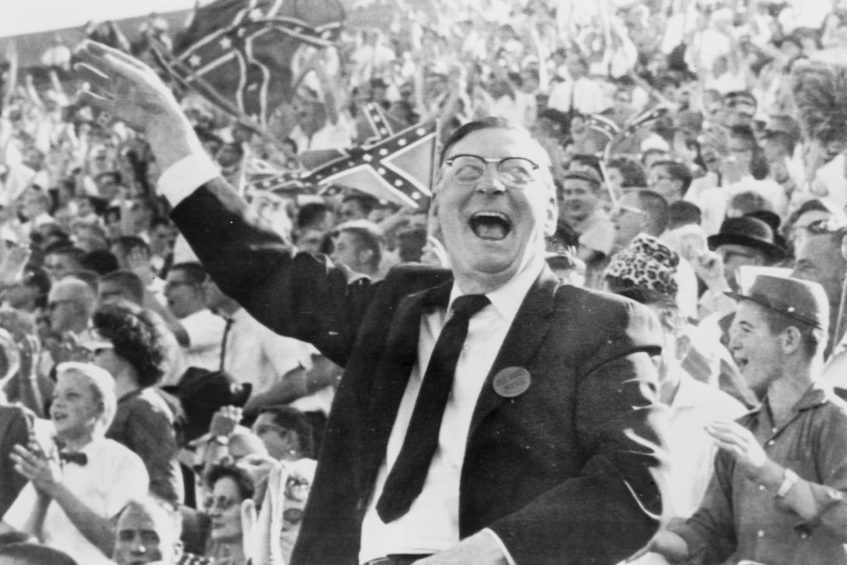 a photo of a man in a suit raising his hand while smiling in front of a crowd waving Confederate flags in a stadium
