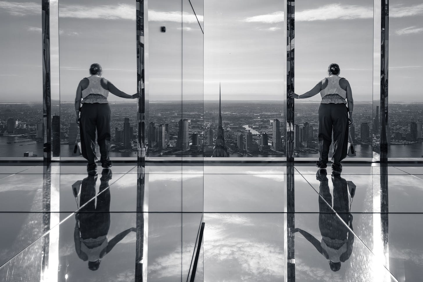 A person leaning on a glass wall doubled by the mirrored panels beside them and doubled again by the mirrored floor, a tall spire standing between the two figures, in black and white. A person leaning on a glass wall doubled by the mirrored panels beside them and doubled again by the mirrored floor, a tall spire standing between the two figures, in black and white.