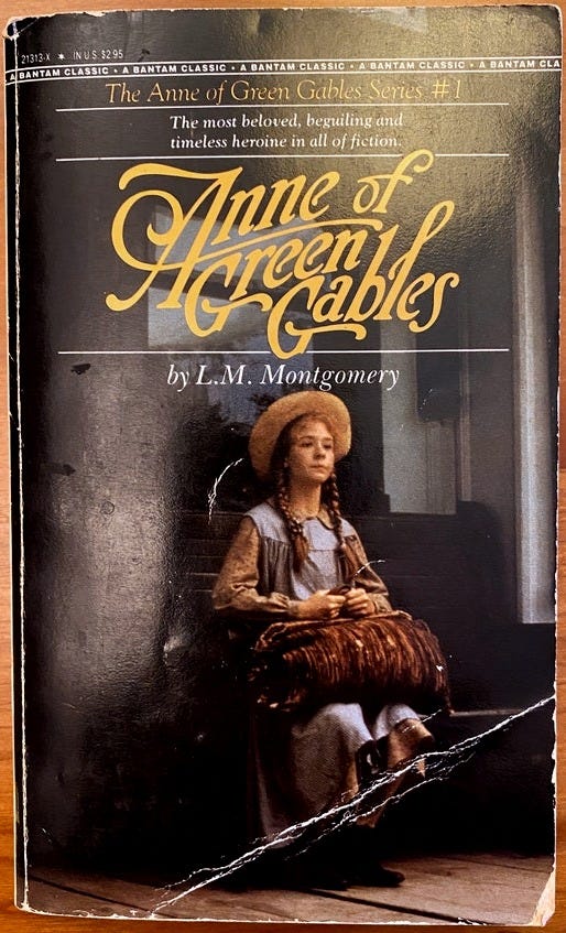 Photograph of a battered paperback copy of the Bantam Classics edition of Anne of Green Gables. On the cover, Megan Follows as Anne (from the 1985) film sits alertly on a bench, clutching a carpet bag. She's wearing a straw hat and has red hair in two braids. The title is above the image in large yellow script.