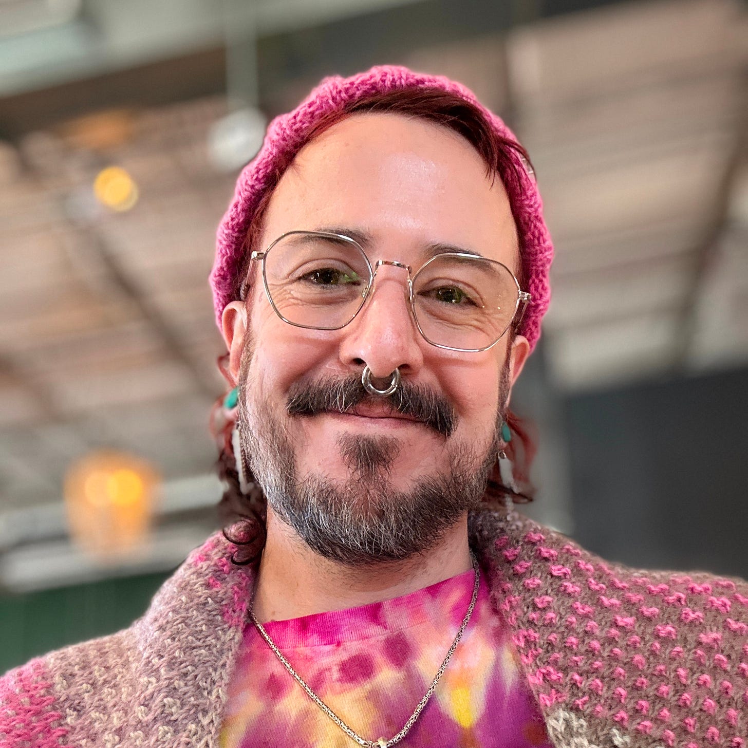 A radical faerie wearing lots of pink, some tie die, wire rimmed glasses and shiny jewelry looks lovingly into the camera.