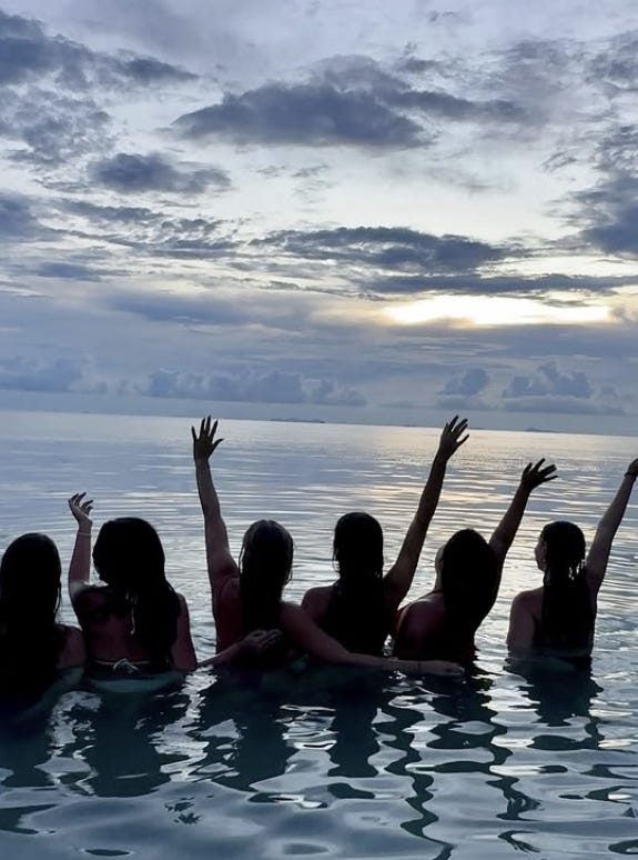 Six people stand in the ocean looking out at the sunset, silhouetted against the horizon with their arms raised in celebration.