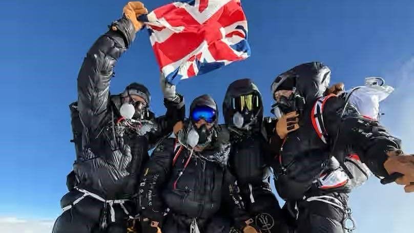 Four climbers standing together, dressed in climbing gear and masks, with one holding up a Union Jack flag Four climbers standing together, dressed in climbing gear and masks, with one holding up a Union Jack flag