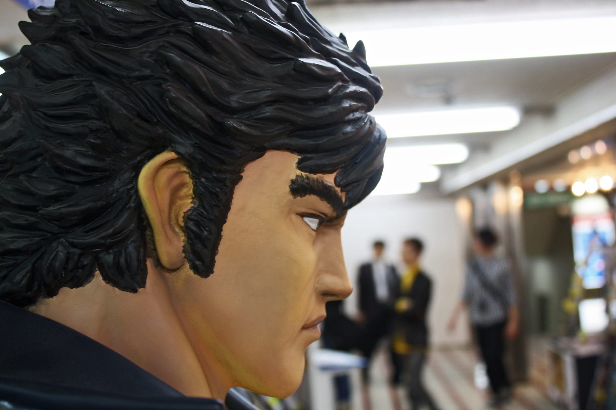 A close-up side view of a large statue of Kenshiro from Fist of the North Star, displayed indoors with blurred figures of people in the background.