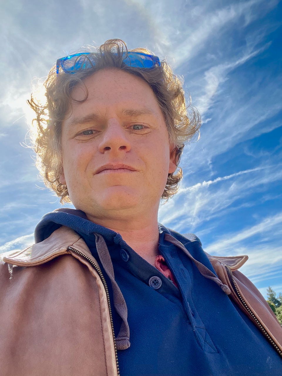 A headshot of Will Fertman, taken selfie-style from below with a blue sky in the background. Will is a white man with curly reddish hair, wearing blue glasses on top of his head and a dark blue shirt and leather jacket.