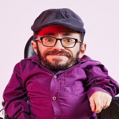 Raul Krauthausen trägt ein magentafarbenes Hemd und eine graue Mütze, auf seinem Kopf sitzt ein grauer Hut. Er hat einen Bart und trägt eine Brille.