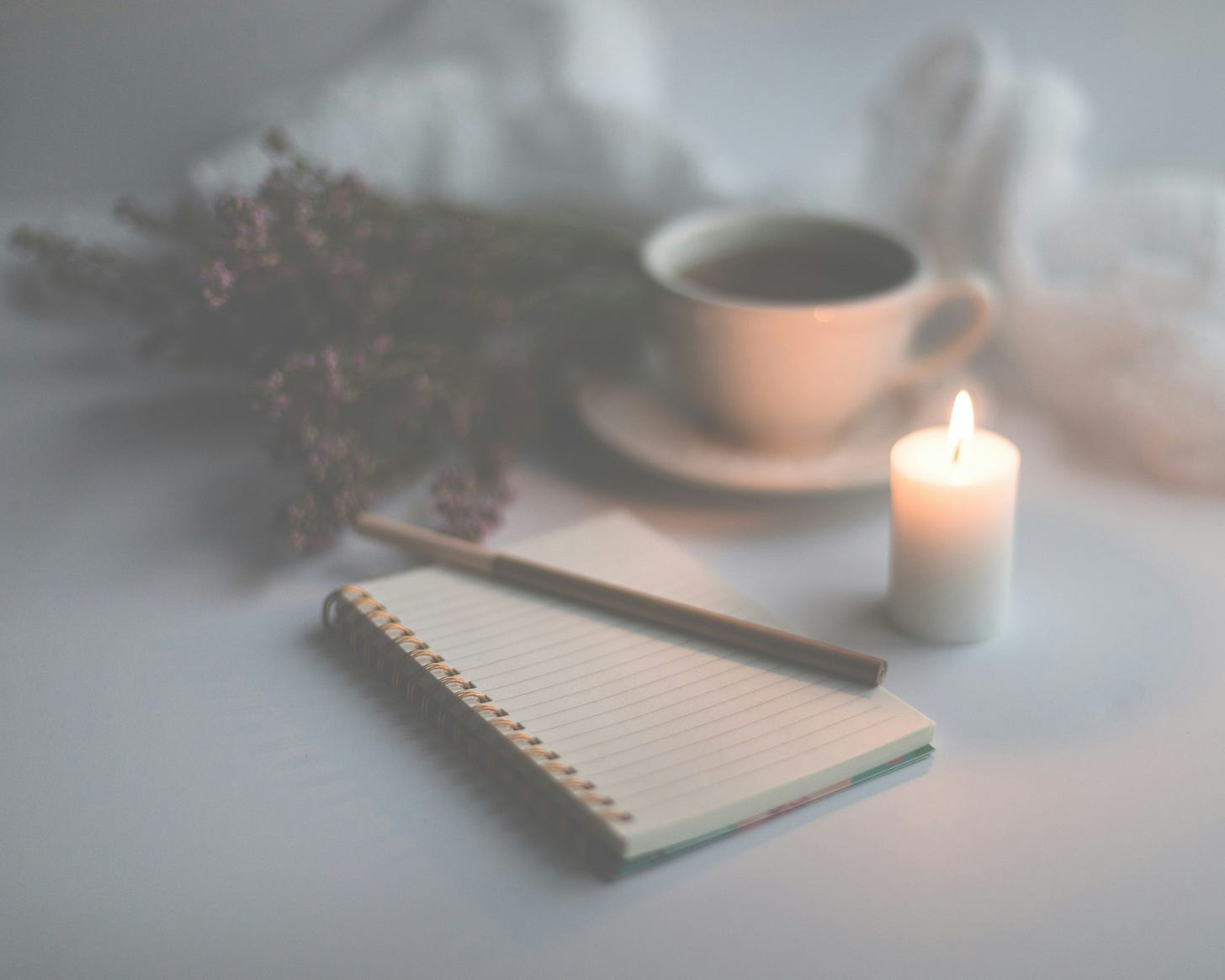 A calming scene with a lit white candle, an open lined notebook with a pen resting on it, a cup of tea or coffee on a saucer, and a bundle of dried lavender in soft focus, all set against a muted, gentle background.