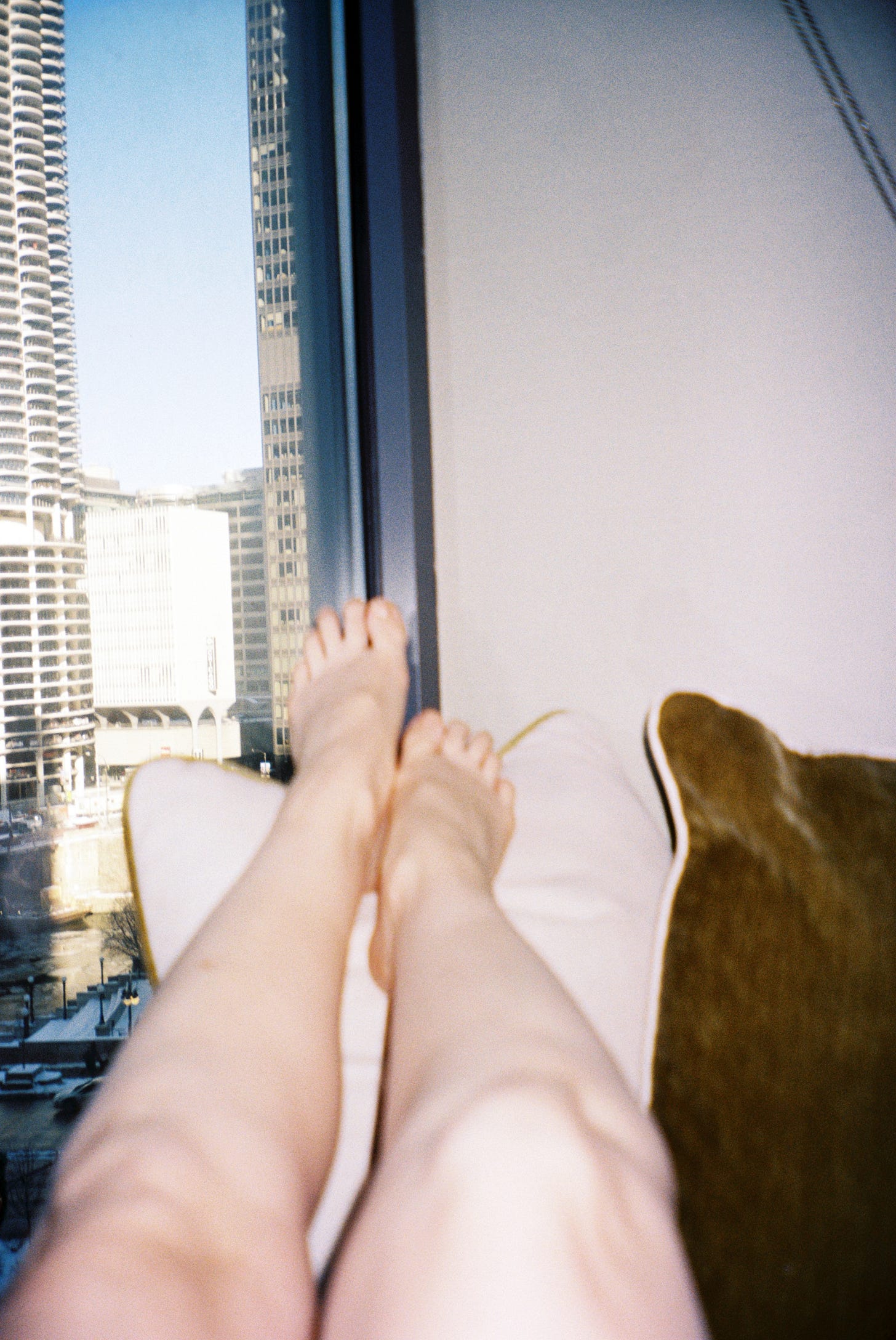 An out of focus film photo of Evyn Silver's legs resting against beige and green pillows. On the right side of the image is a beige wall, and on the left side is a window looking out onto bright blue sky and tall skyscrapers.