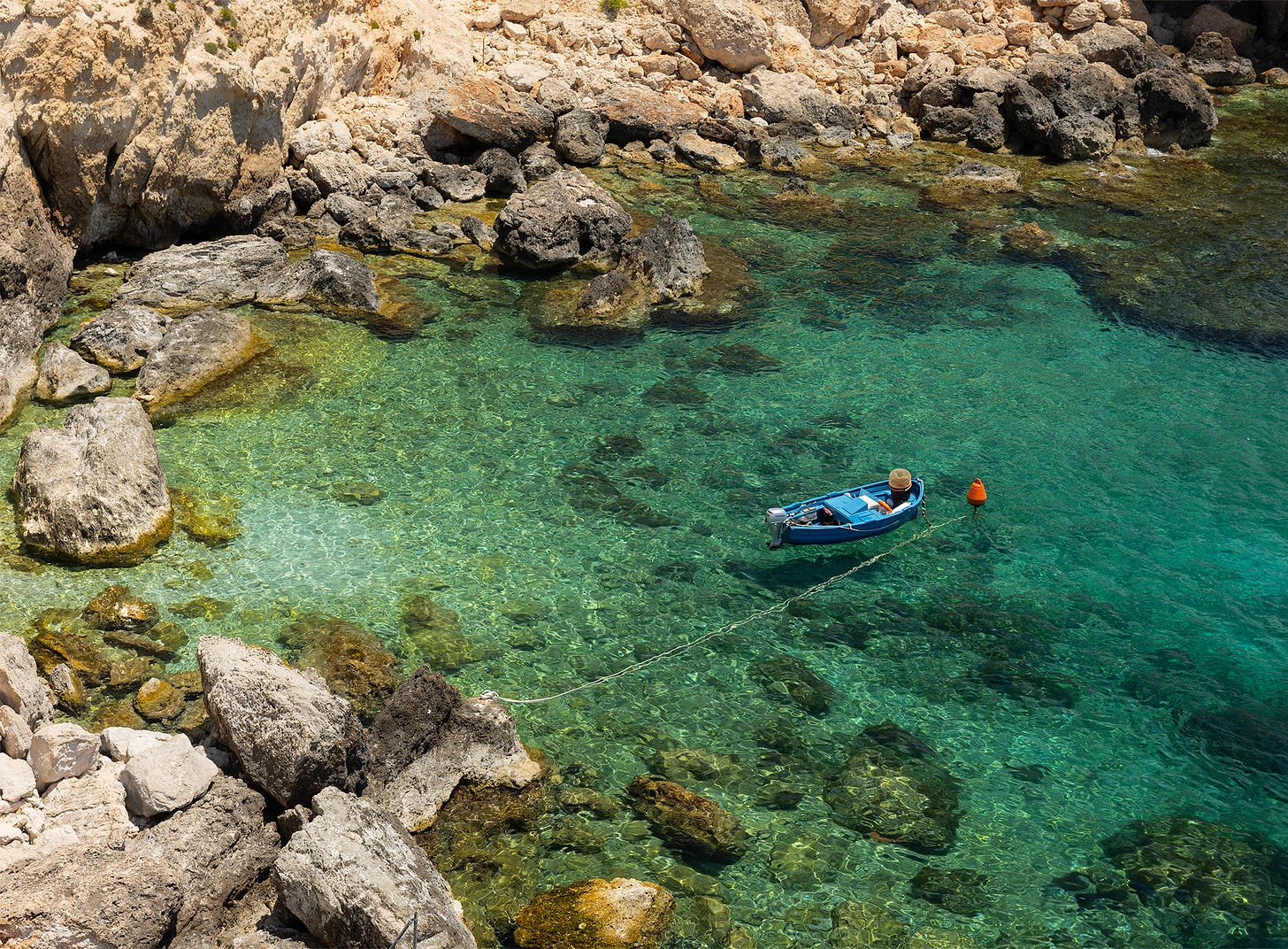 Small blue boat floating in the crystal-clear turquoise waters of Għar Lapsi, Malta, surrounded by rocky shoreline and submerged stones.