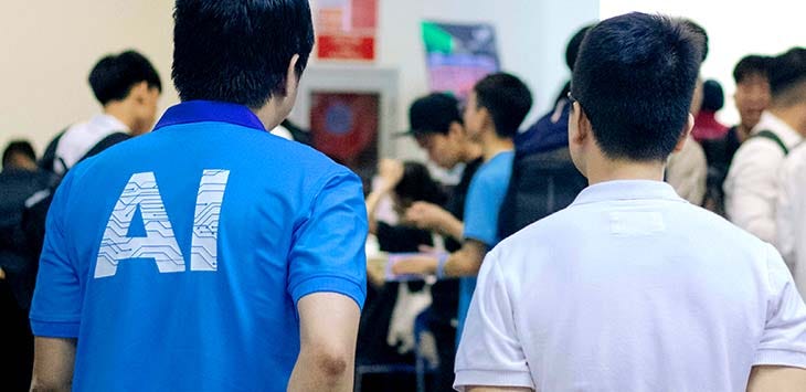 Photo taken from behind two men, one of whom is wearing a t-shirt emblazoned with the capital letters A.I. on the back.