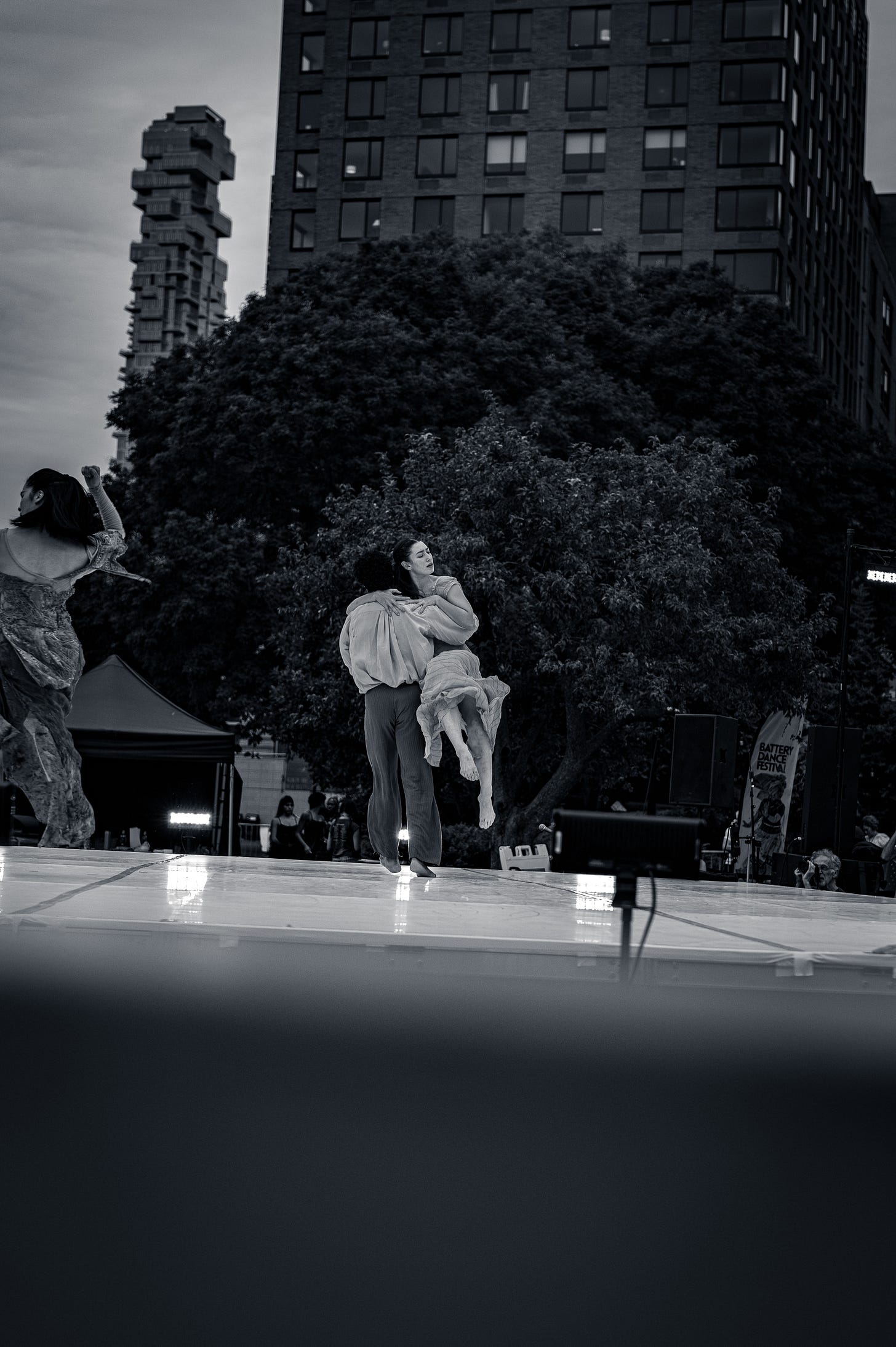 A male dancer holds a female dancer mid-performance on an outdoor stage, with trees and tall buildings in the background. A male dancer holds a female dancer mid-performance on an outdoor stage, with trees and tall buildings in the background.