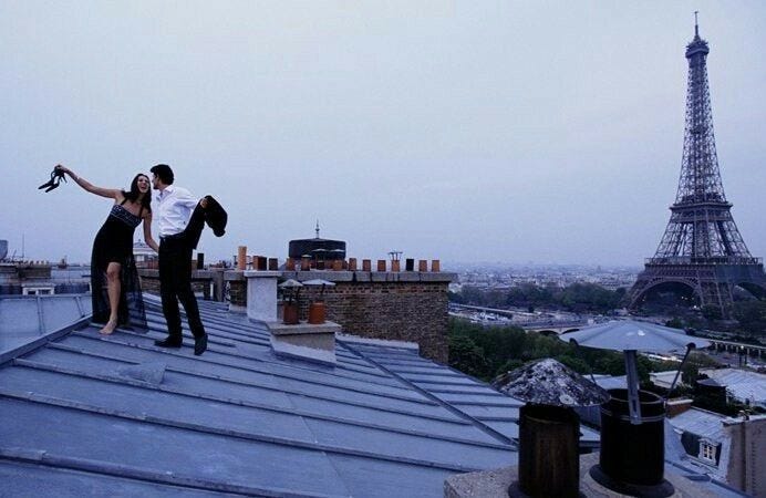 This may contain: two people standing on the roof of a building in front of the eiffel tower