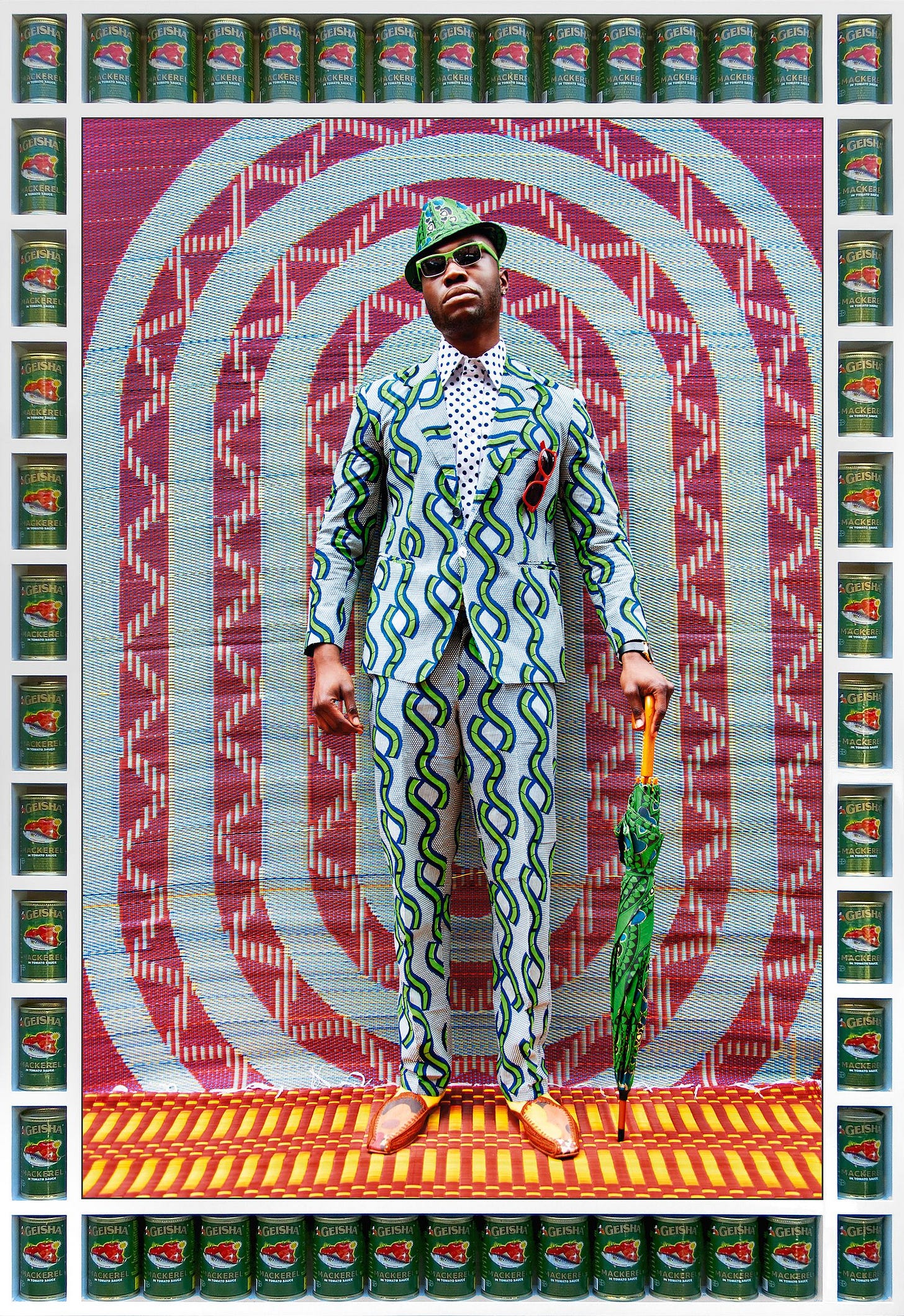 A young Black man wearing sunglasses standing and looking directly at us, dressed in a light blue and green African wax print suit against a backdrop that is equally vibrant with another African wax print cloth which is mainly dark red and turquoise. This portrait by Hassan Hajjaj has a custom made frame that encases tins of Geisha mackerel making the overall effect of the clashing colours somewhat dizzying. But that is the point.