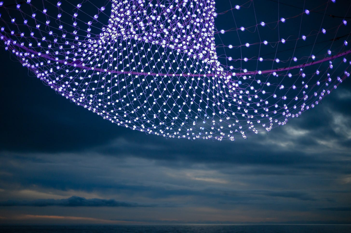 A collection of colourful LED lights hang in front of a dusk sky looking out to sea.
