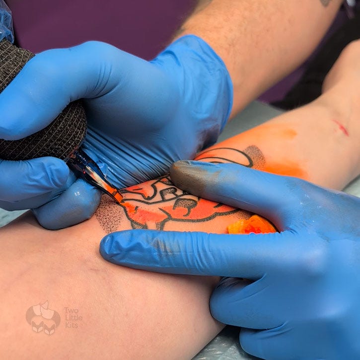 A work-in-progress photo of a tattoo of two foxes on a caucasion woman's inner arm. The style is cartoonish with bold outlines and dotted shading. The tattoo artist is working on the orange colouring. A work-in-progress photo of a tattoo of two foxes on a caucasion woman's inner arm. The style is cartoonish with bold outlines and dotted shading. The tattoo artist is working on the orange colouring.