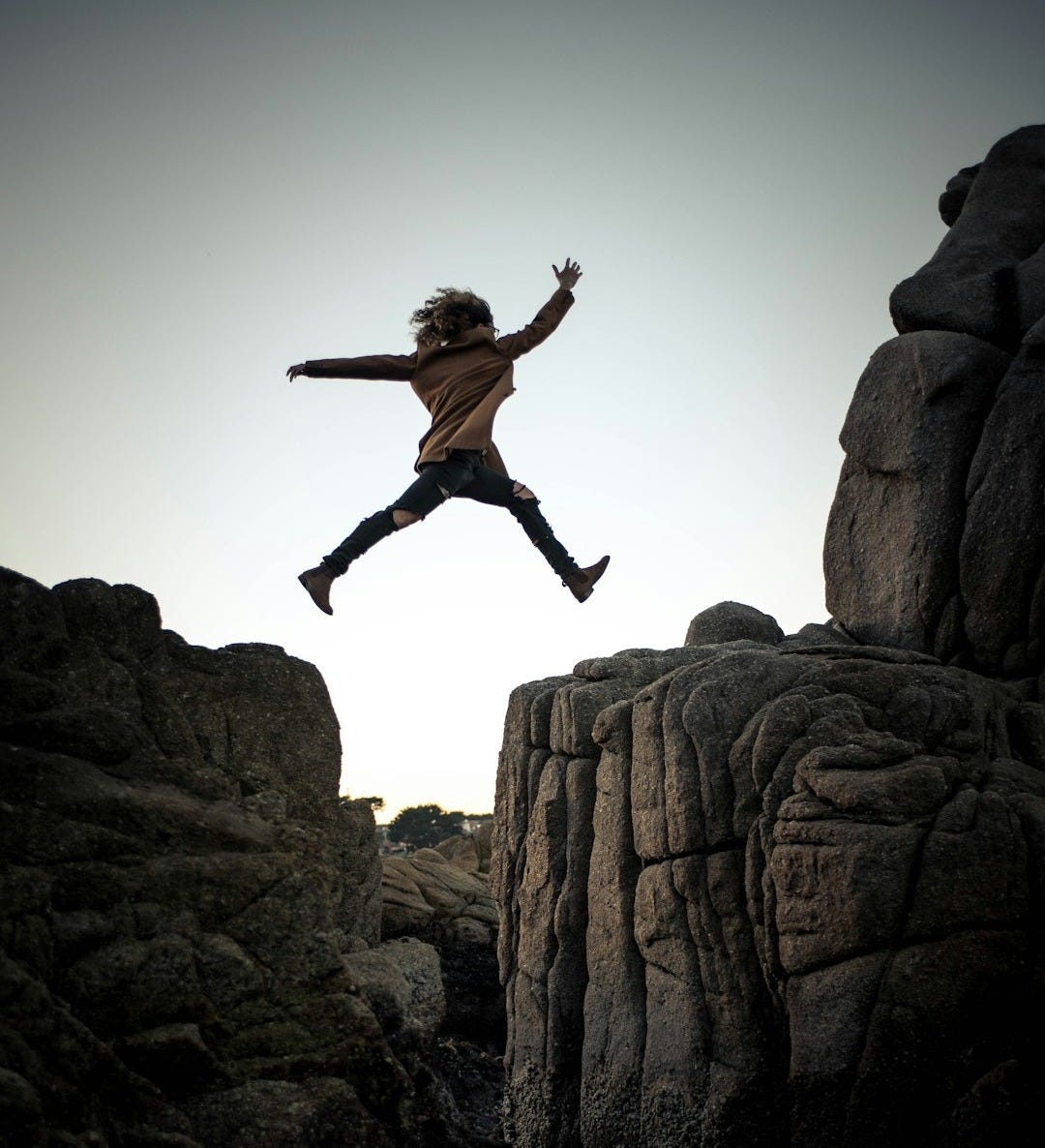 person jumping on big rock under gray and white sky during daytime
