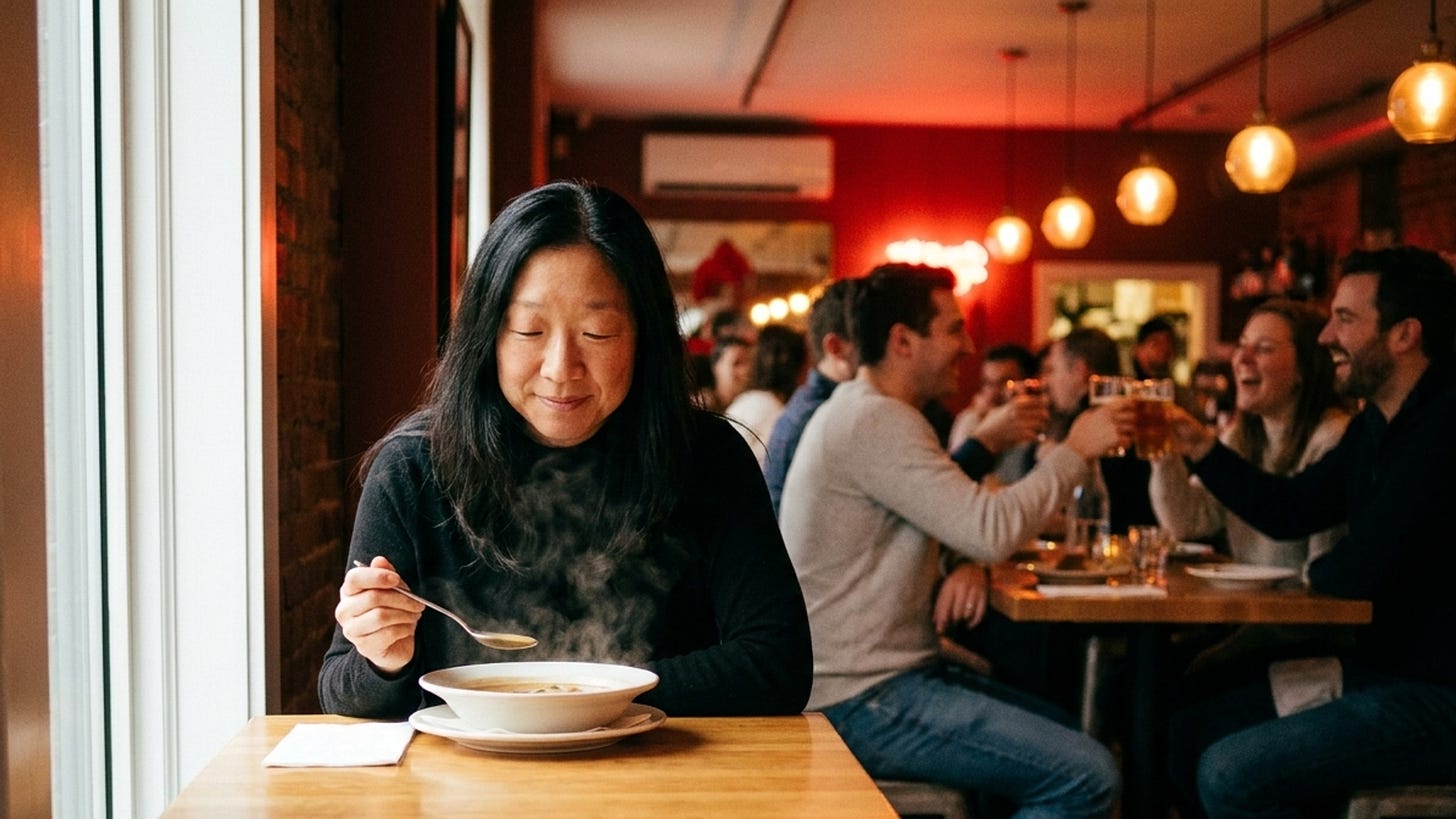 A composed Asian woman in her 40s with long dark hair sits alone at a small table in a lively upscale restaurant, gently holding a spoon over a bowl of piping hot soup with visible steam rising. Soft natural light illuminates her face. Around her, groups of friends celebrate at other tables—laughter, toasting glasses, animated conversations. She is not reading, not on her phone, just present with the soup. The composition contrasts her chosen stillness with the surrounding social energy.