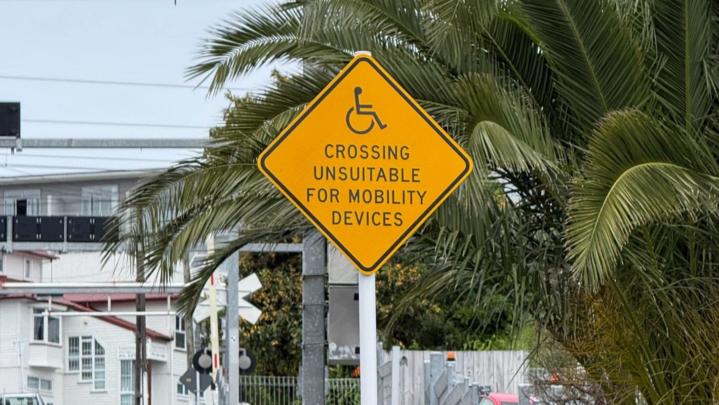 A diamond-shaped yellow sign with a wheelchair symbol and the words “crossing unsuitable for mobility devices” on an urban residential road. 