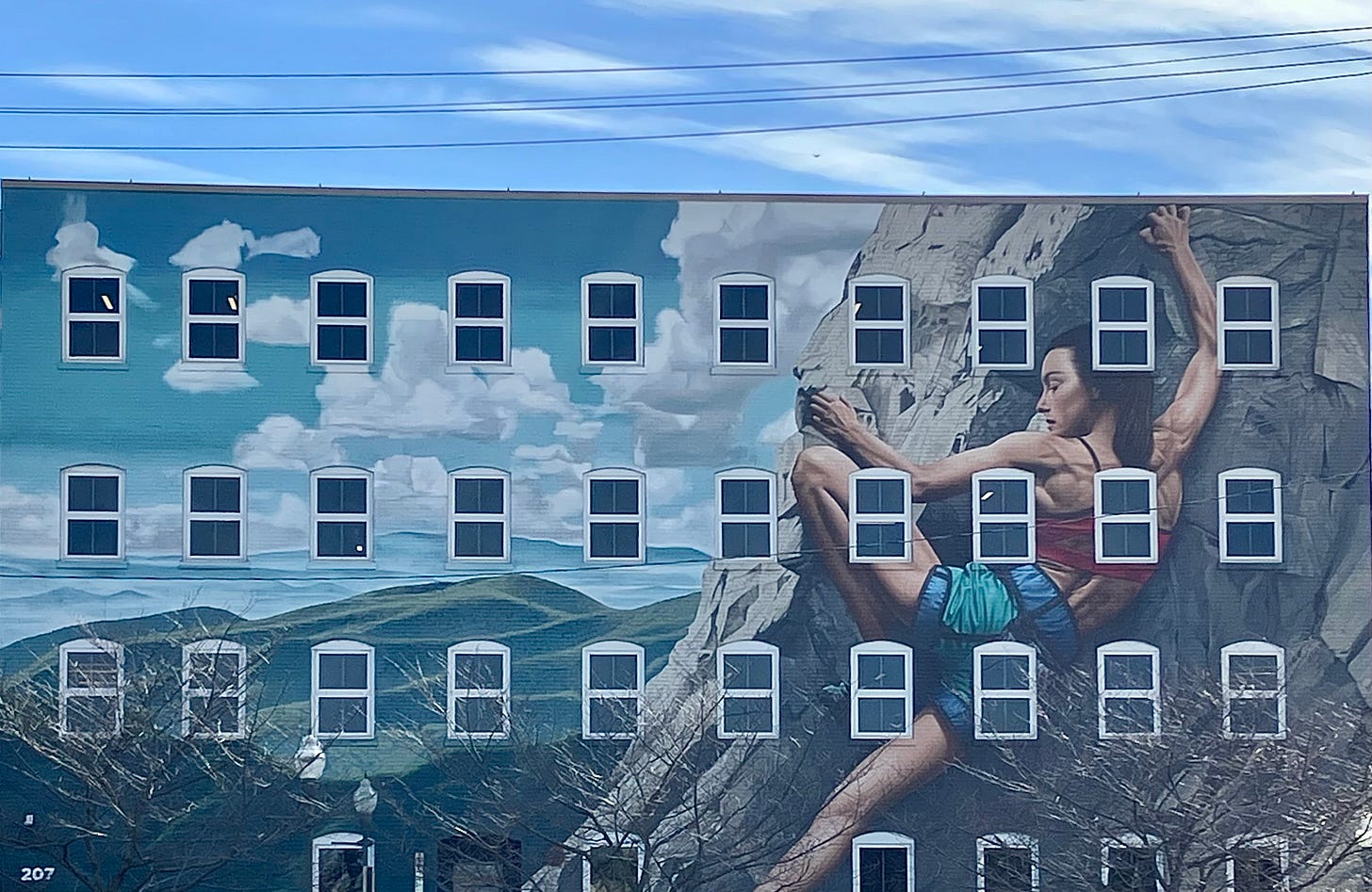 The photo is of a mural on a large building with many windows. It shows a blue sky with puffy clouds over a green mountains. To the right, a woman in climbing gear is holding onto a rock face with her fingers and toes.