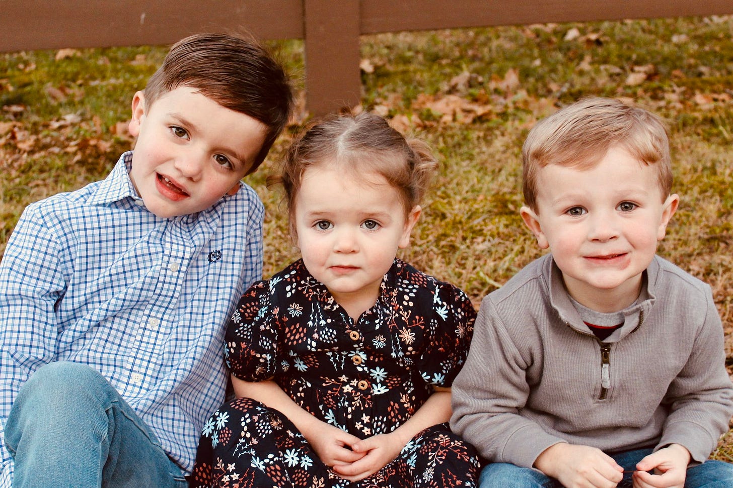 Three young children sit in the grass in front of a brown fence. Three young children sit in the grass in front of a brown fence.