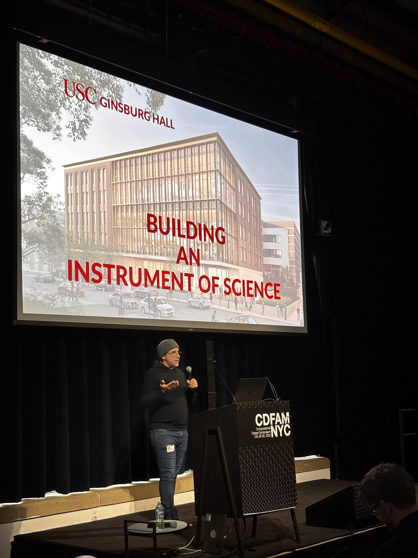 A speaker at a podium presents at a conference about the USC Ginsburg Hall, emphasizing its role as an 'Instrument of Science' with an image of the building projected behind.