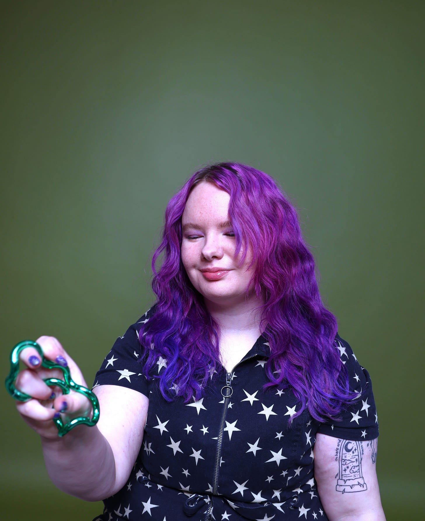 Charli is a white person with purple hair wearing a starry jumpsuit against a green background