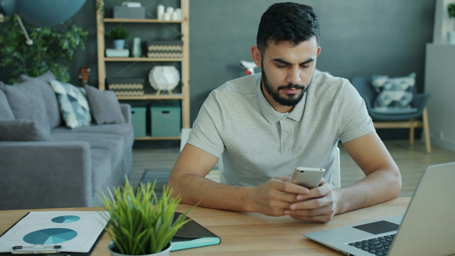 A man at a desk in a living room looks down at his phone, with an open laptop beside him and printed charts and a plant on the table. A man at a desk in a living room looks down at his phone, with an open laptop beside him and printed charts and a plant on the table.