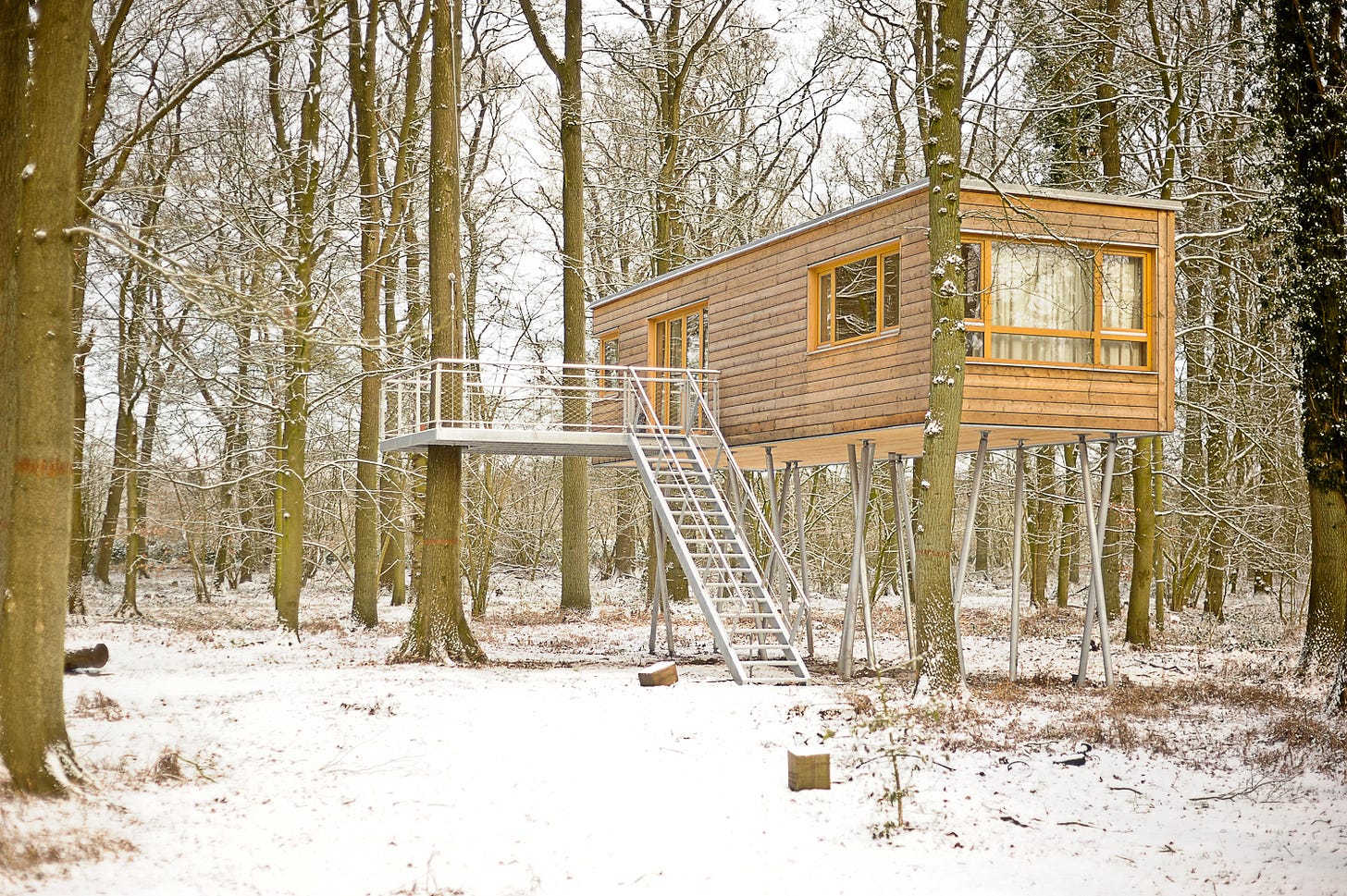 A long wooden-slatted cabin on stilts with a staircase and balcony surrounded by trees. There is snow on the ground.