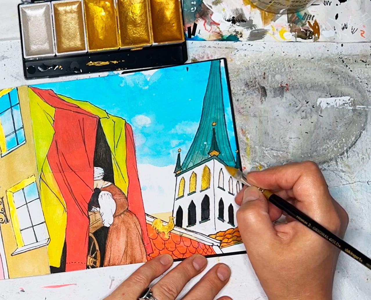 Photo showing a close-up of panels in a sketchbook, and my hand holding a paintbrush loaded with metallic gold paint, filling in a drawing of the arched windows of a medieval church steeple