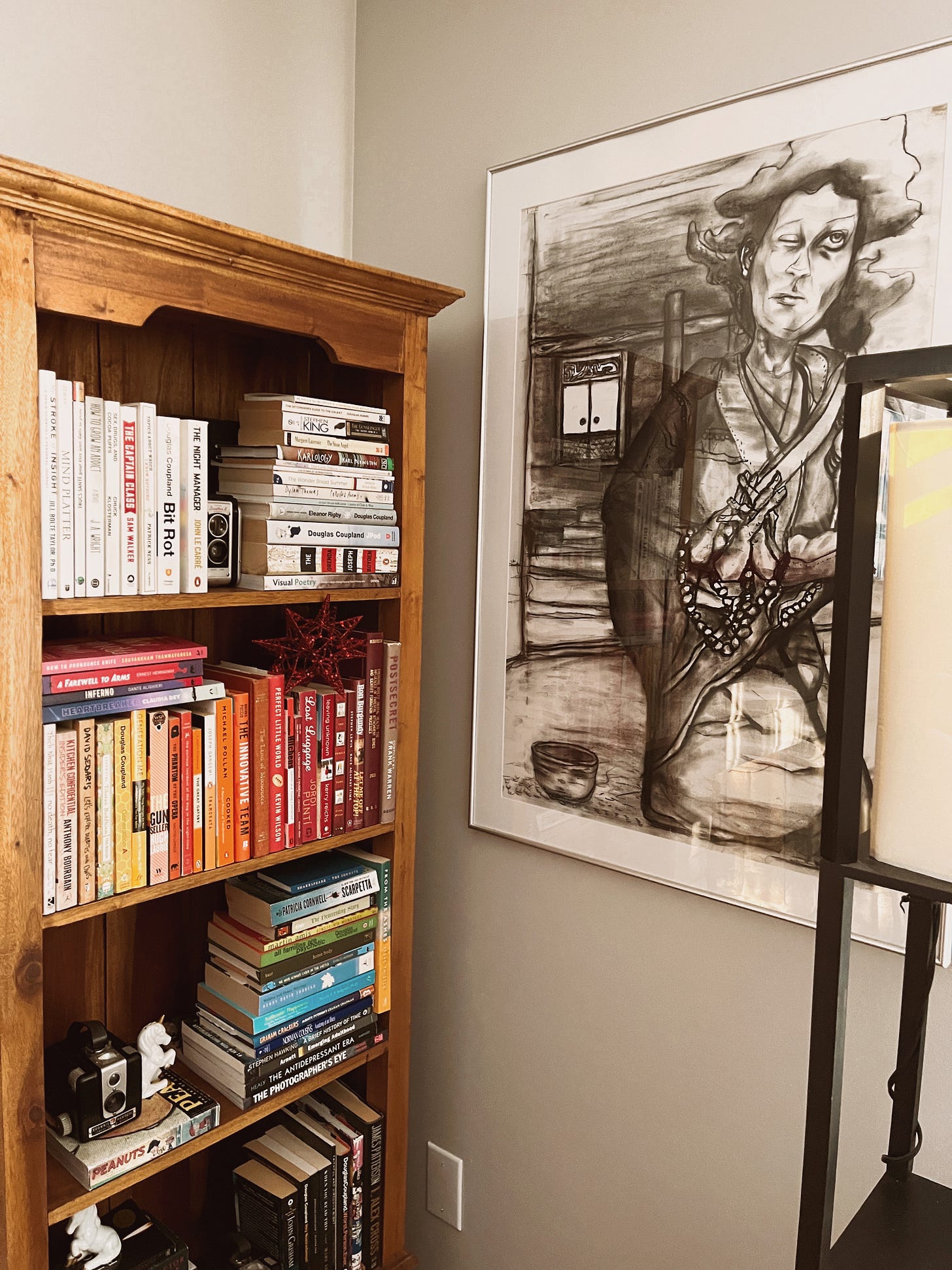 Large charcoal drawing of a woman clutching prayer beads and looking askance at the heavens, one eye open. Next to the picture is a bookshelf where I've organized my books by colour of the covers. Large charcoal drawing of a woman clutching prayer beads and looking askance at the heavens, one eye open. Next to the picture is a bookshelf where I've organized my books by colour of the covers.