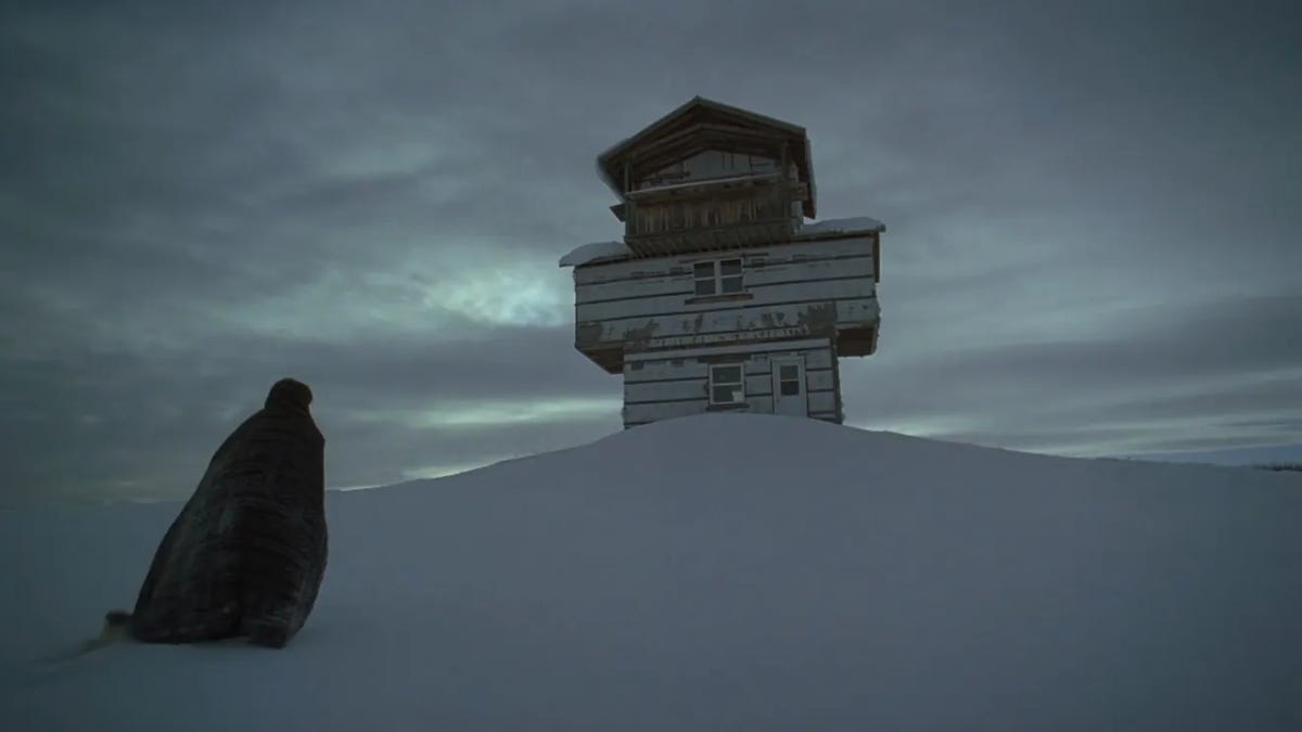 A figure in a robe walks up a snowy hill to a run-down cabin under a cloudy sky.