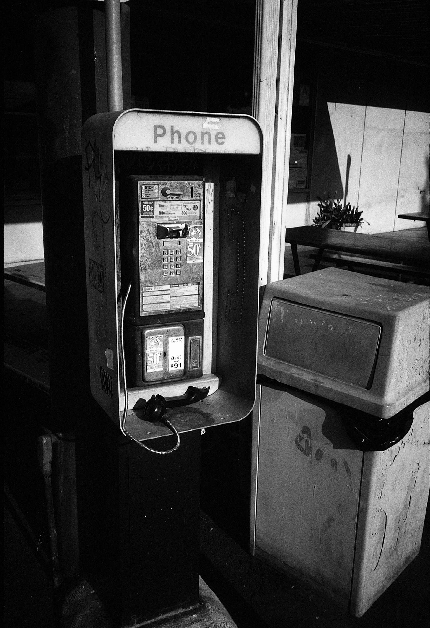 Black and white photo of an old, beat-up phone booth with a broken phone next to a trash can