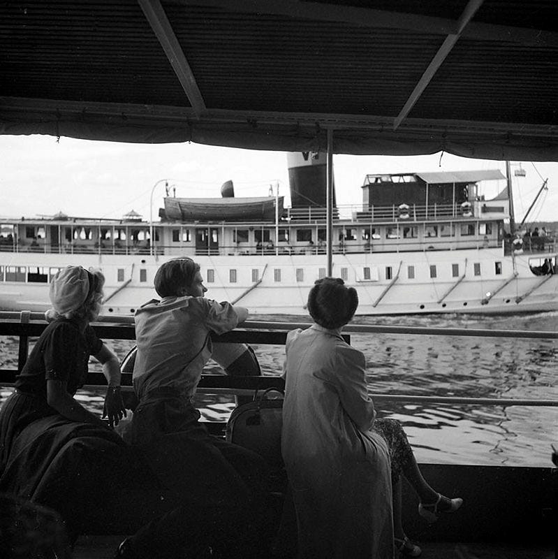 Black and white image of three people sitting on a boat, looking across at a small passenger ferry in 1940s Stockholm