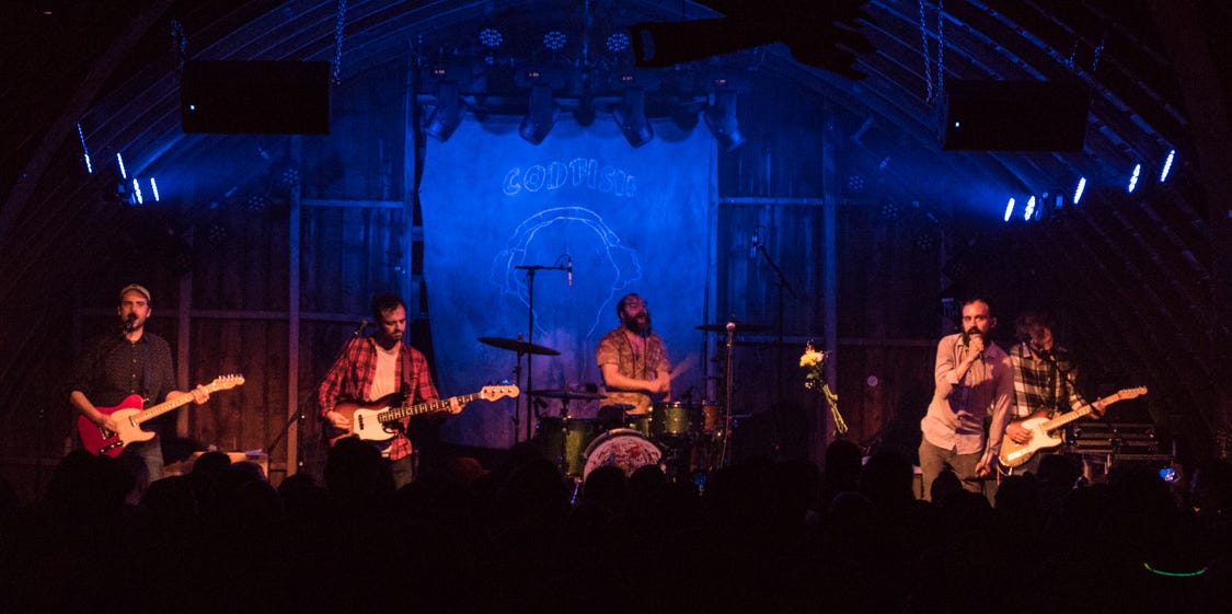 Four men performing on stage. From right to left: man in a dark shirt with an electric guitar, man in a red flannel with an electric bass guitar, man in a tan shirt behind a drum kit, man in a pink shirt with a microphone in his hand, and a man black and white shirt with an electric guitar. Four men performing on stage. From right to left: man in a dark shirt with an electric guitar, man in a red flannel with an electric bass guitar, man in a tan shirt behind a drum kit, man in a pink shirt with a microphone in his hand, and a man black and white shirt with an electric guitar.