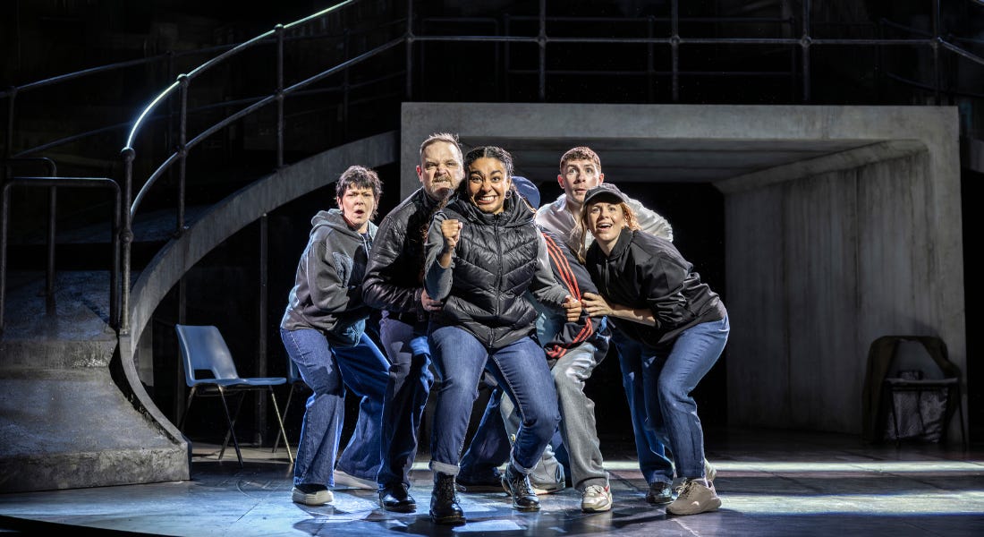 A group of 6 people wearing blue jeans and dark jackets crowd together. They look out with excited, expectant expressions. The woman at the front clenches her fist in victory.