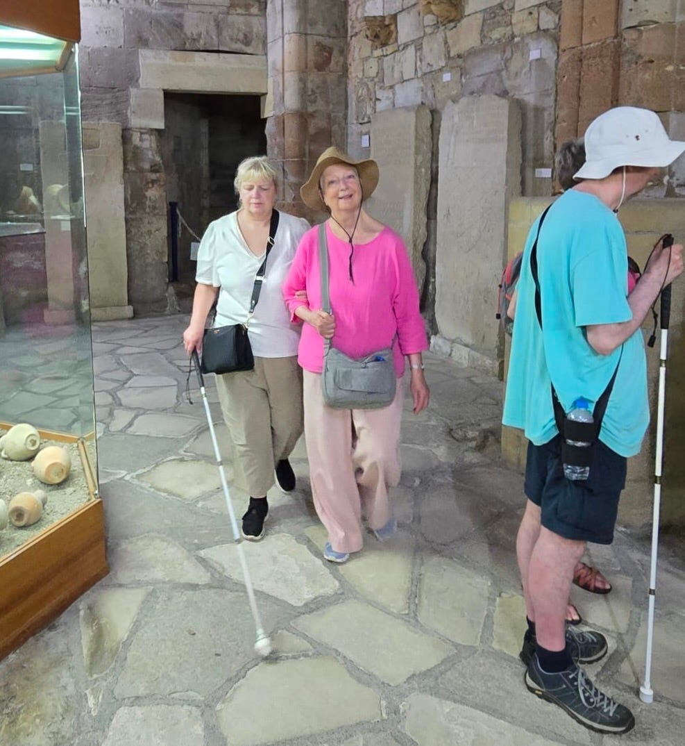 Two women walk towards the person holding the camera. They are in an old castle building with stone floors and walls. There is a display cabinet to the left of the picture with pots behind glass.