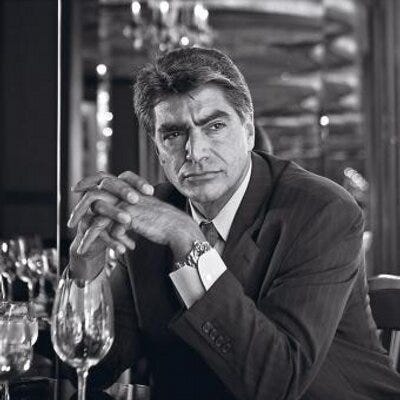 Wearing a suit, Mark Rossini leans over an empty glass at a restaurant table