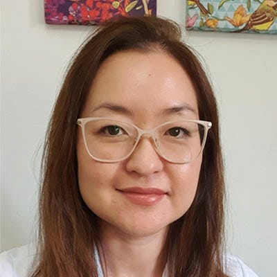 A imagem mostra um retrato de uma mulher adulta, enquadrada do peito para cima, olhando diretamente para a câmera. Ela tem pele clara, rosto oval e expressão tranquila, com um leve sorriso. Seus cabelos são lisos, de cor castanho médio, na altura dos ombros, repartidos levemente de lado. Ela usa óculos de armação clara e transparente, com lentes grandes.